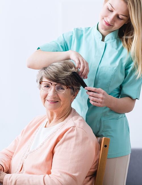 A carer in a teal uniform gently brushing an older woman's hair at home