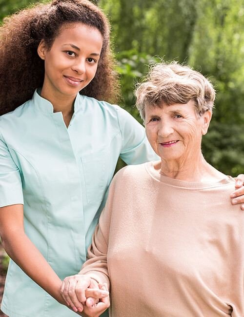 An Obatos carer in a mint-green uniform gently holding the hand of an older woman outdoors