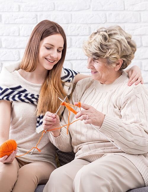 A carer and an older woman sitting close together, knitting and laughing in a bright living room