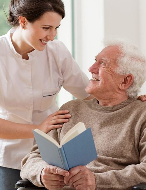 A carer reading a book with a smiling older man seated in his living room