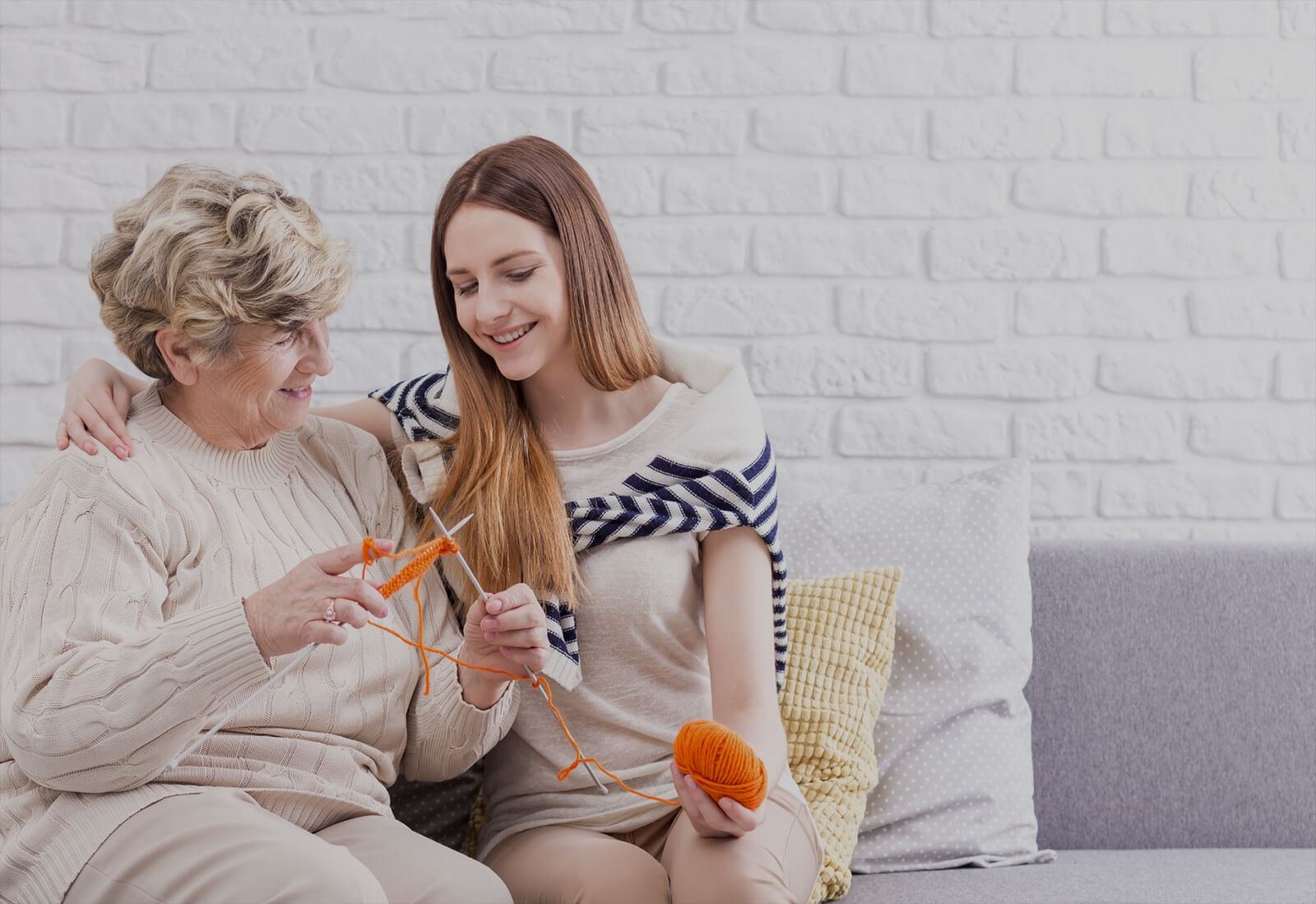 A younger woman and an older woman sitting close together on a sofa, laughing while knitting
